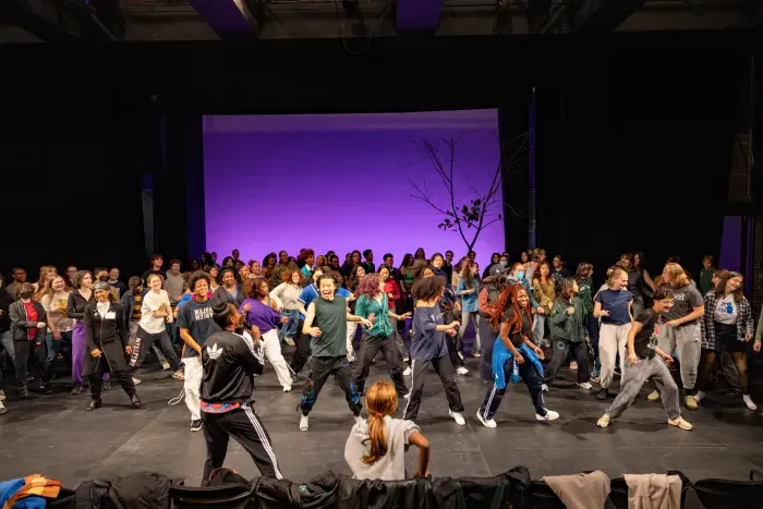 A large group of students on a stage in front of a purple backdrop, dancing as a group