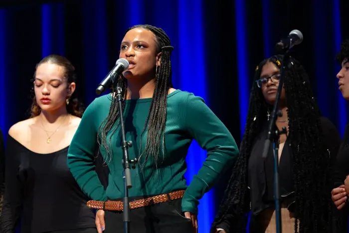 A student in a long-sleeved green shirt stands with her hands on her hips, singing into a microphone